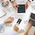 Manos en una mesa con portátil, tableta, móvil y bocetos de gráficos